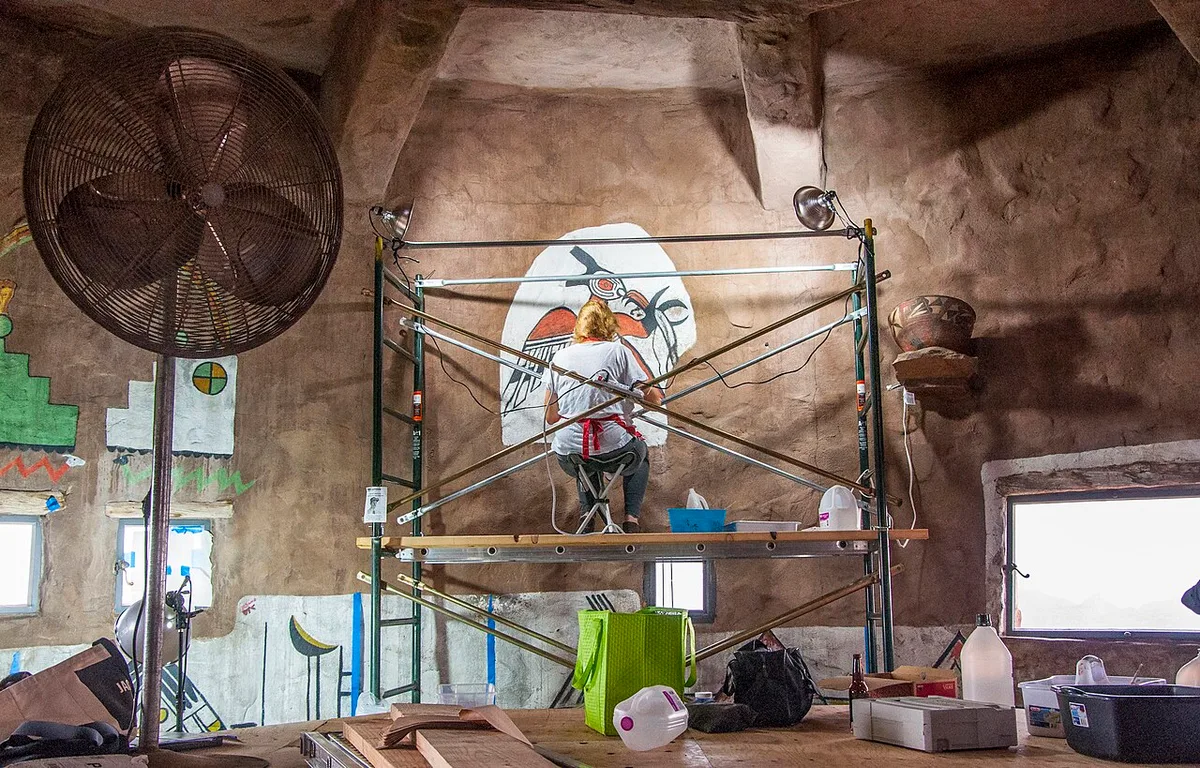 Een restaurator werkt vanaf een steiger aan een beschilderd wandpaneel, met gereedschap, jerrycans en ventilatoren op de vloer eromheen.