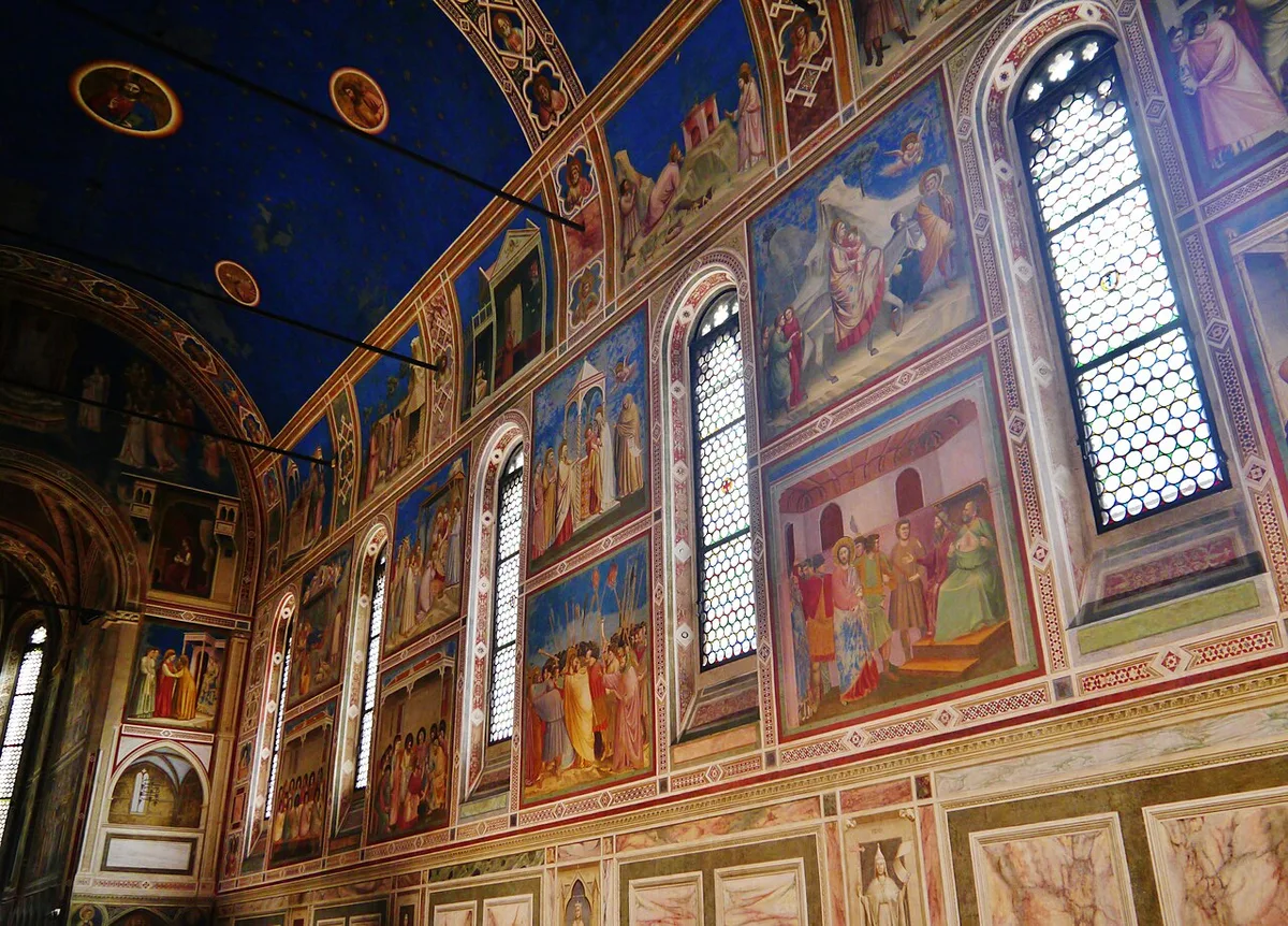 Het schip van de Scrovegni-kapel in Padua: een blauw tongewelf met sterren boven wanden vol fresco's in rijen, met hoge spitsboogramen in de rechterwand.