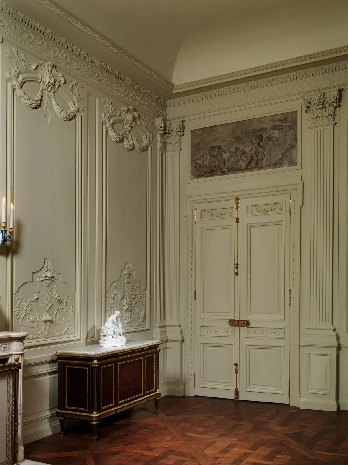 Een lichte, wit geschilderde houten wandbetimmering met gesneden cartouches, een reliëf boven de deur en dubbele paneeldeuren met vergulde beslag.