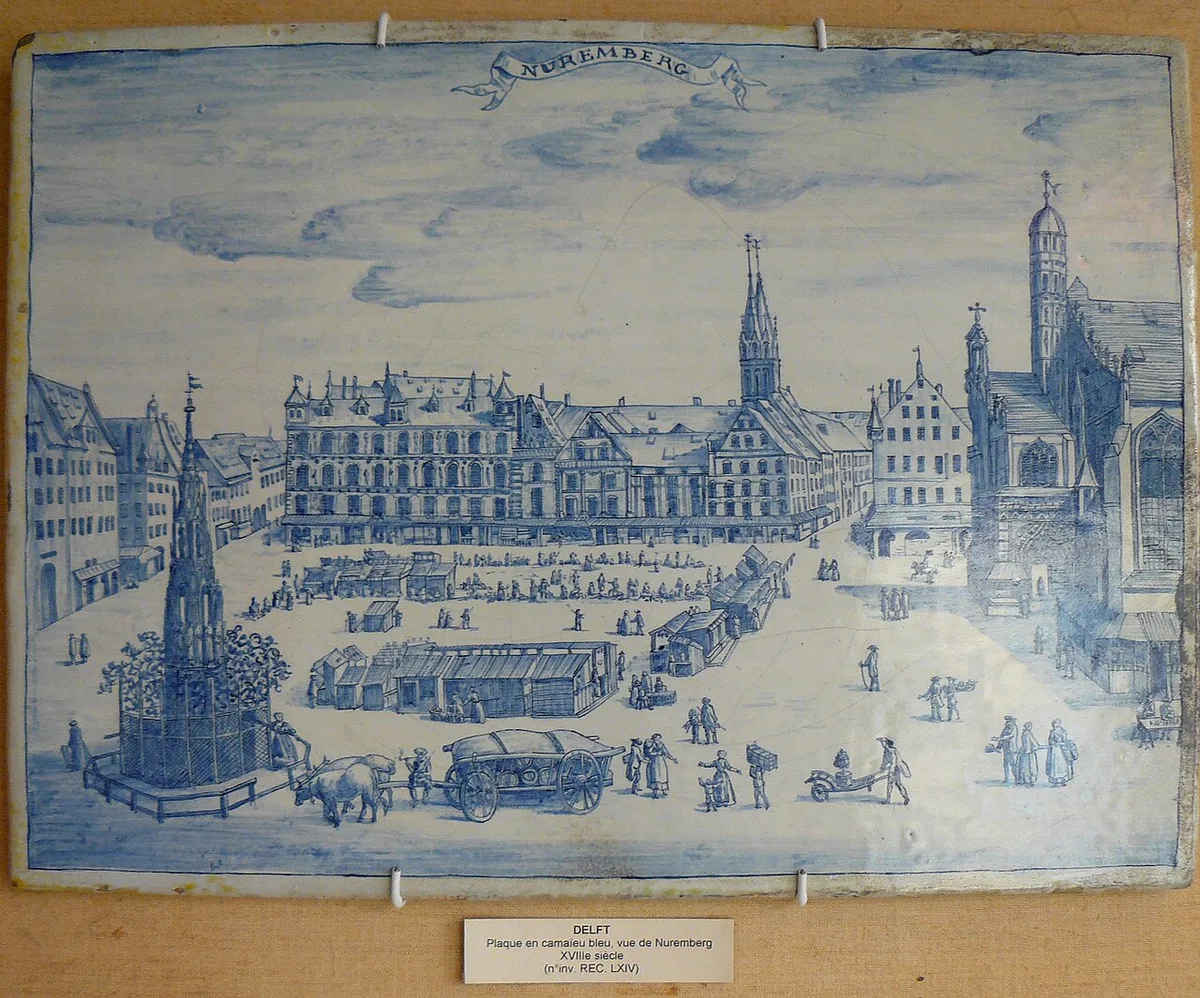 Een Delfts tegeltableau in blauw op wit met een stadsgezicht van de markt van Neurenberg: gevels, een fontein, marktkramen en kleine figuren.